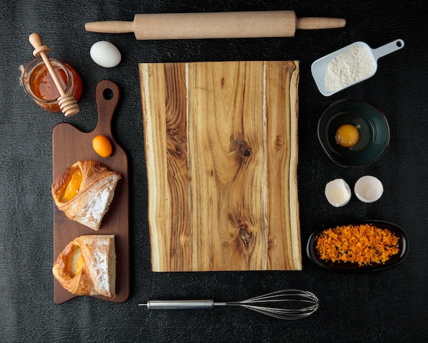 Vista dall'alto del tagliere con croissant tagliato marmellata di mele cotogne uovo scorza d'arancia tuorlo d'uovo guscio d'uovo mattarello su sfondo nero