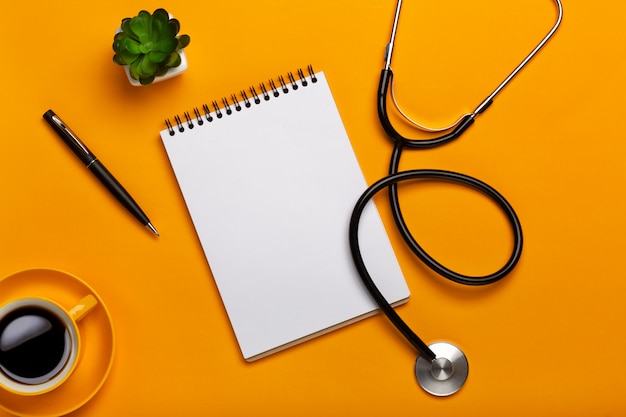 Vista dall&#39;alto del tavolo di un medico con blocco note e penna stetoscopio, tastiera, prescrizione e pillole, una tazza di caffè su uno sfondo giallo