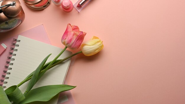 Vista dall'alto dell'area di lavoro piatta creativa femminile rosa con fiori, taccuino e cosmetici sul tavolo