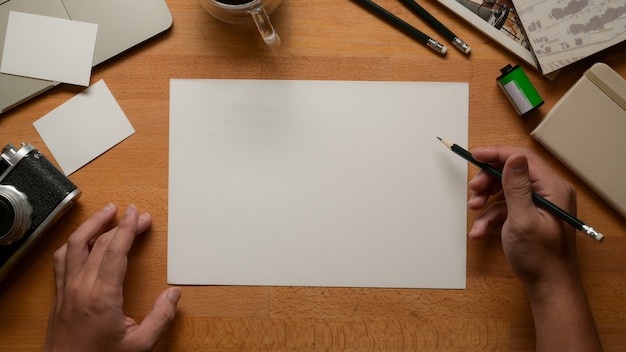 Vista dall'alto dell'artista maschio che lavora con carta da disegno in bianco, matite, fotocamera e forniture sul piano di lavoro