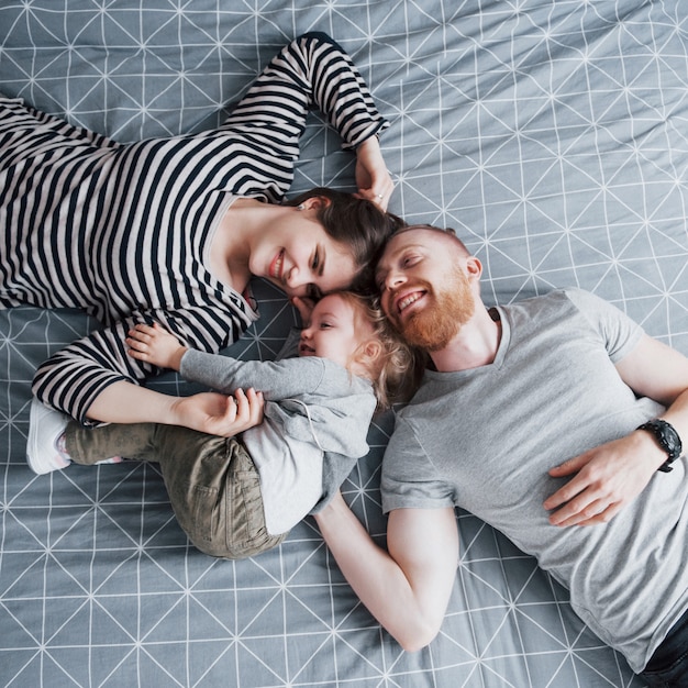 Vista dall'alto della bellissima giovane madre, padre e la loro figlia guardando la fotocamera e sorridente mentre giaceva sul letto testa a testa