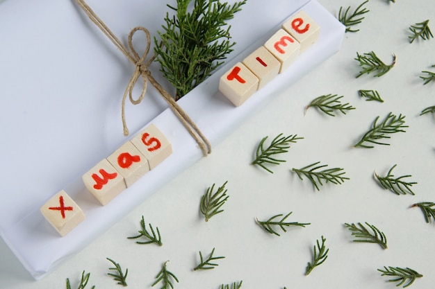 Vista dall'alto della confezione regalo bianca artigianale con testo "X mas time" (lettere in legno) e rami di abete fresco su sfondo bianco, celebrazione stagionale e concetto ecologico