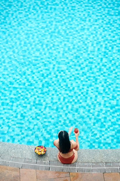 Vista dall'alto della donna dai capelli neri in costume da bagno che beve cocktail e mangia frutta mentre si rilassa...