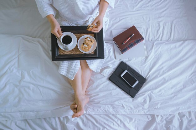 Vista dall'alto della donna in accappatoio bianco che beve caffè con colazione dopo il lavoro online sul letto