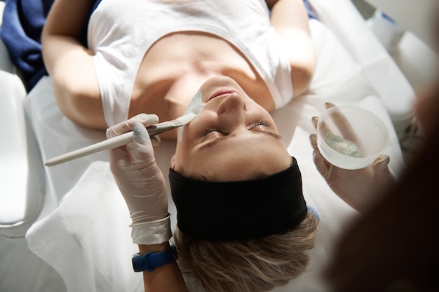 Vista dall'alto della giovane donna che si rilassa su un lettino da massaggio durante la procedura di beauty spa e medico cosmetologo che applica una maschera di alginato sul viso. Concetto di cura della pelle professionale nel salone SPA