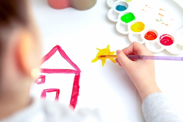 Vista dall'alto della piccola mano del bambino che disegna la casa e il sole con gli acquerelli. Istruzione e concetto di scuola.