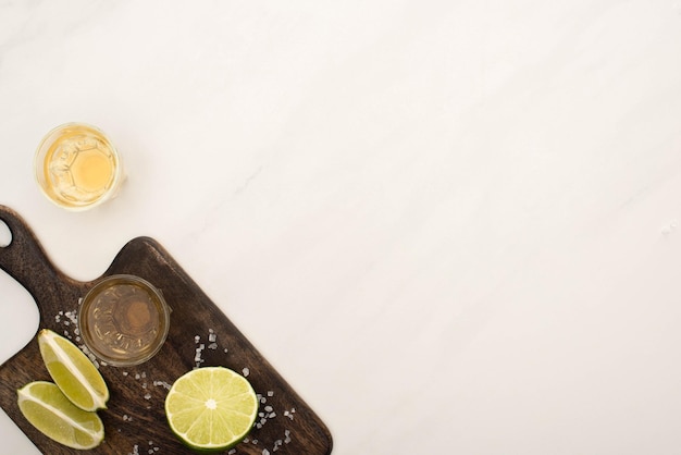 Vista dall'alto della tequila dorata con sale di calce su tagliere di legno su superficie di marmo bianco