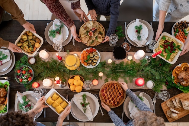 Vista dall'alto delle mani dei membri della famiglia che tengono piatti con cibo e dolci fatti in casa mentre servono la tavola festiva prima della cena di Natale