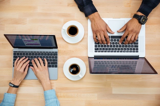 Vista dall'alto delle mani dell'uomo e della donna che lavorano con due laptop e bevono caffè su un tavolo di legno