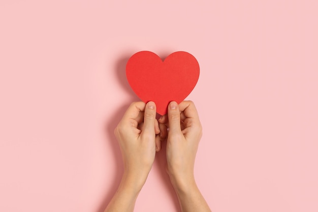 Vista dall'alto delle mani di donna che tengono carta bianca saluto o carta di invito su sfondo rosa pastello con forma di cuore