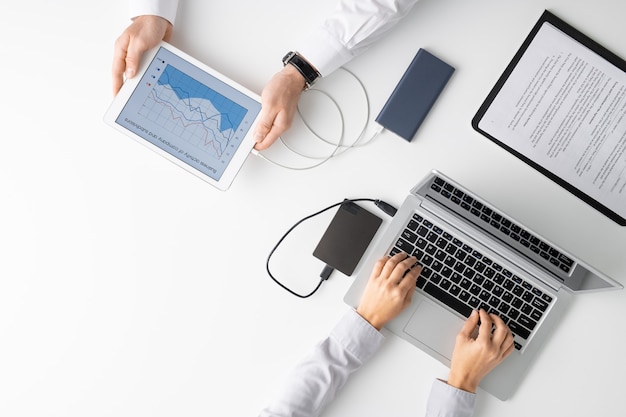 Vista dall'alto delle mani di due medici che utilizzano gadget mobili mentre uno di loro analizza i grafici e il suo collega digitando sul laptop