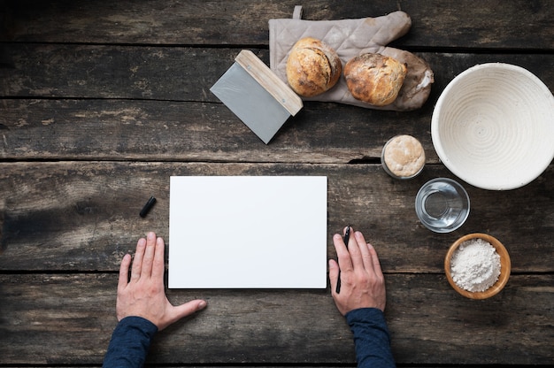 Vista dall'alto delle mani maschile in procinto di scrivere una ricetta per un pane a lievitazione naturale sano fatto in casa su un pezzo di carta bianco con tutti gli ingredienti disposti intorno ad esso sulla scrivania in legno rustico.