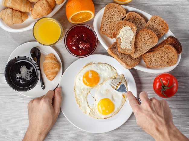 Vista dall'alto delle mani maschili che tengono piatto con uova fritte e forchetta al tavolo con una tazza di caffè nero, croissant, ciotola con marmellata di fragole, pane sul piatto, succo d'arancia fresco