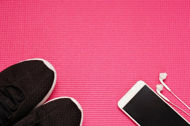 Vista dall'alto dello smartphone con auricolare e sneaker su tappetino Yoga rosso Fitness e concetto sano