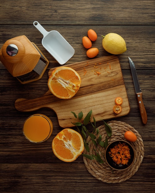 Vista dall'alto di agrumi come limone kumquat arancia tagliata a metà sul tagliere con scorza d'arancia succo d'arancia coltello misura cucchiaio e grattugia su fondo di legno