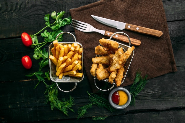 Vista dall'alto di appetitose patatine fritte e pollo fritto in cestini di metallo su un tavolo di legno