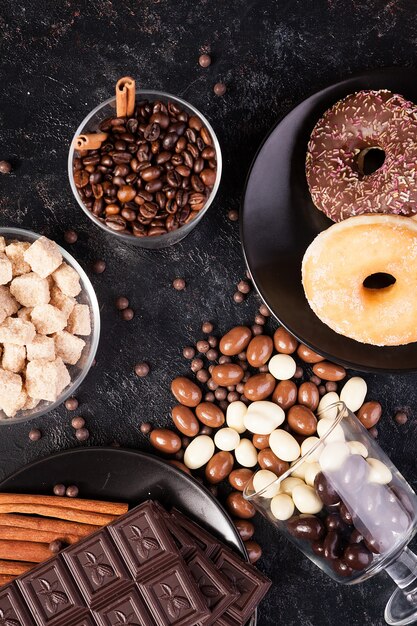 Vista dall'alto di arachidi nel cioccolato, versate sul bordo scuro, accanto a tavolette di cioccolato, ciambelle, zucchero di canna e chicchi di caffè. Foto in studio