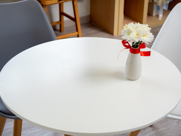 Vista dall'alto di bellissimi fiori bianchi e gialli in fiore in vaso di ceramica ornato con decorazione a nastro rosso su tavolo rotondo in legno con ampio spazio vuoto per la pubblicità del prodotto