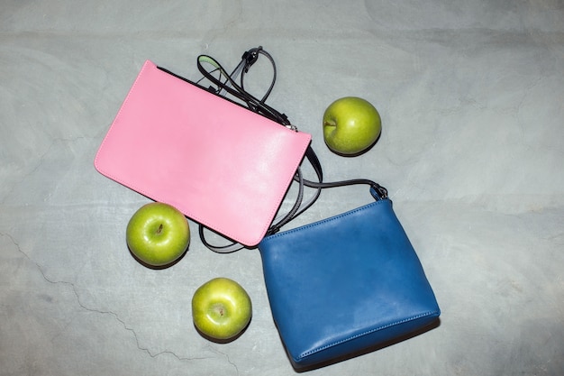 Vista dall'alto di borse in pelle blu e rosa alla moda disposte sul tavolo con mele verdi fresche che mostrano il concetto di materiali eco-compatibili