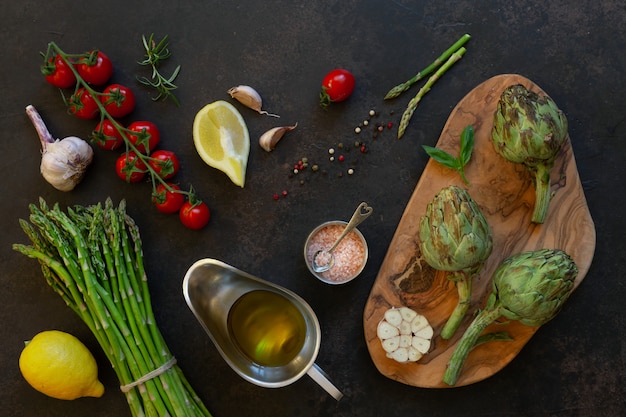 Vista dall&#39;alto di carciofo fresco e mazzo di asparagi verdi con olio d&#39;oliva, pomodori, limone e aglio sul tavolo