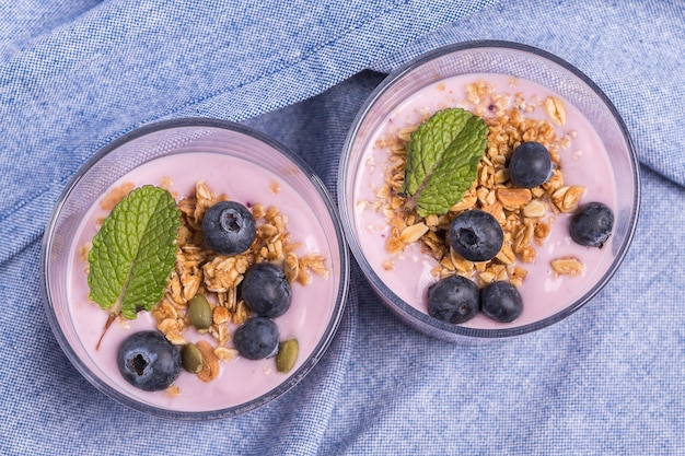 Vista dall'alto di due bicchieri di yogurt alla fragola con mirtilli e cereali su una tovaglia blu