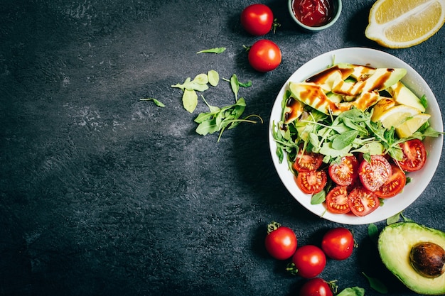 Vista dall'alto di insalata di pomodori rucola avocado limone in piatto bianco su sfondo nero