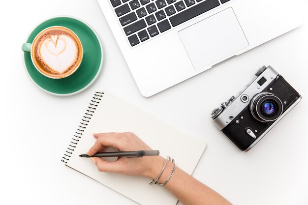 Vista dall'alto di laptop, vecchia macchina fotografica, tazza di caffè e scrittura a mano di una donna su un taccuino su sfondo bianco