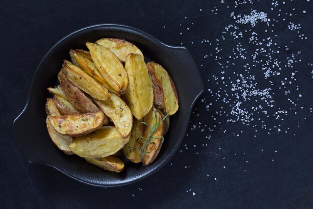 Vista dall'alto di patate arrosto rustiche con ramo di rosmarino su piatto nero su sfondo nero