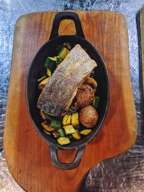 Vista dall'alto di pesce al forno, verdure, patate con erbe aromatiche in una padella di ghisa in cucina. bar