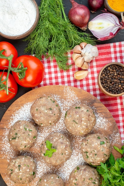 Vista dall'alto di polpette di carne crude con verde in un piatto marrone e verdure fresche farina pepe verde su asciugamano rosso su sfondo nero