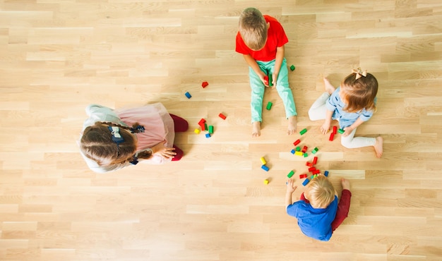 Vista dall'alto di quattro adorabili bambini che giocano su un pavimento con blocchi di legno. Ritratto di bambini seduti a piedi nudi che giocano in tranquilla cooperazione