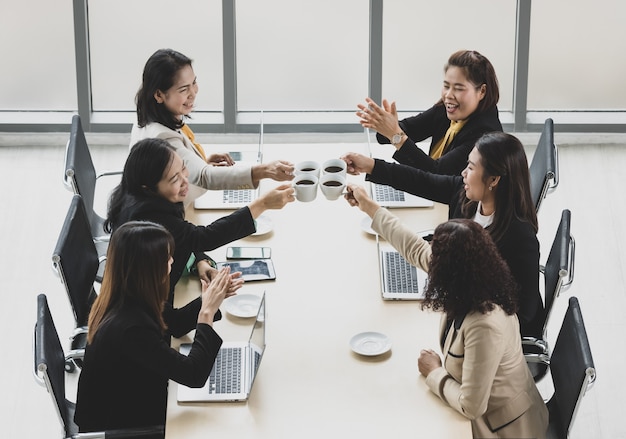 Vista dall'alto di sei donne d'affari sedute insieme attorno a un tavolo da conferenza in legno, tifo e tintinnanti tazza da caffè insieme a laptop e tablet sul tavolo in ufficio. Concetto per incontro di lavoro.