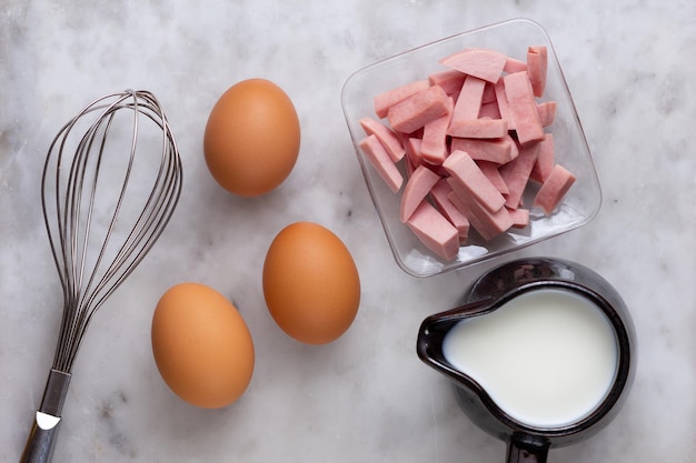 Vista dall'alto di tre uova marroni crude, farina, latte e salsicce tagliate come ingredienti di una frittata sulla superficie di marmo