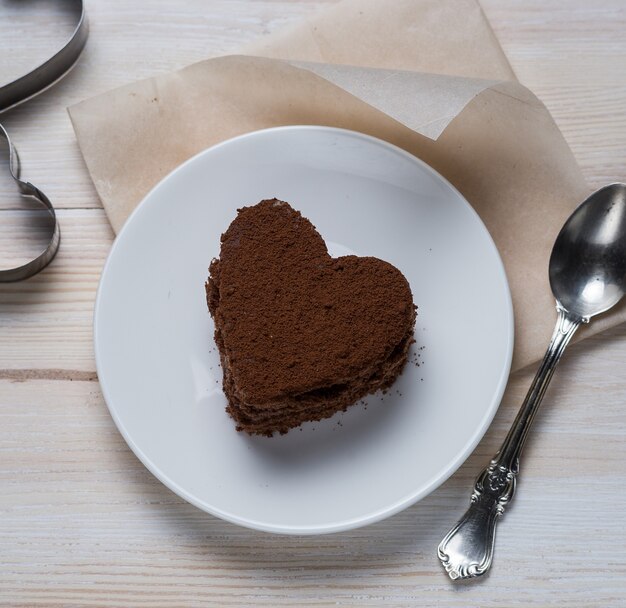 Vista dall'alto di un dolce al cioccolato a forma di cuore sdraiato su un piattino accanto a un cucchiaio d'argento e forme di ferro su uno sfondo di legno bianco.