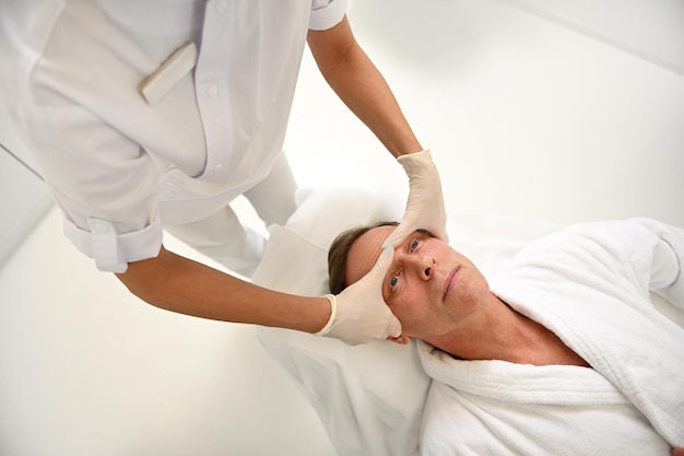 Vista dall'alto di un'estetista irriconoscibile che esegue un massaggio professionale anti-età levigante per il lifting del viso della fronte dell'uomo maturo. Cura della pelle e concetto di procedure di bellezza maschile presso wellness spa