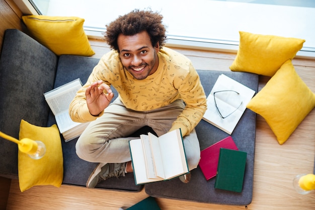 Vista dall'alto di un giovane afroamericano felice che legge il libro e mostra il segno ok a casa