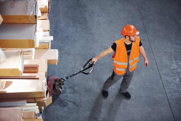 Vista dall'alto di un giovane lavoratore di sesso maschile in uniforme che si trova nel magazzino che spinge il transpallet.