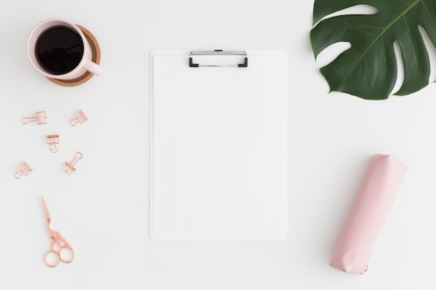 Vista dall'alto di un mockup di appunti bianco con accessori per l'area di lavoro caffè e una foglia di monstera su un tavolo bianco