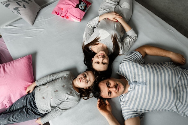 Vista dall'alto di una bambina carina e dei suoi bellissimi giovani genitori che guardano la telecamera e sorridono, sdraiati sul pavimento a casa, Vvew dall'alto della famiglia che riposa sdraiata sul pavimento e guardando la telecamera