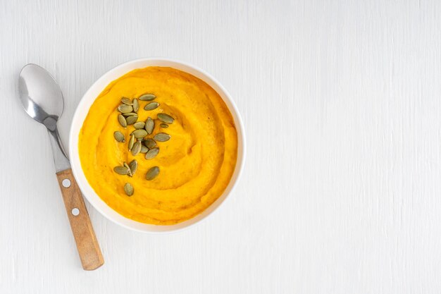 Vista dall'alto di una ciotola di zuppa di zucca cremosa fatta in casa con semi secchi e cucchiaio su un tavolo bianco di legno