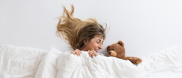 Vista dall'alto di una donna che è sorpresa e guarda un orsacchiotto che dorme sul letto bianco sdraiato sotto la coperta