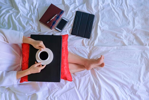 Vista dall'alto di una donna in accappatoio bianco che beve caffè mentre si rilassa dopo aver lavorato online sul suo letto