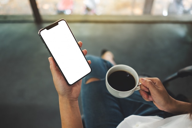 Vista dall'alto di una donna in possesso di un telefono cellulare nero con schermo desktop bianco bianco con una tazza di caffè