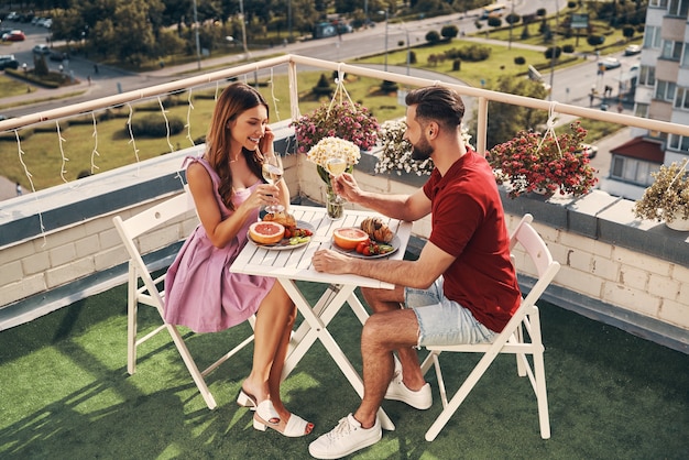 Vista dall'alto di una giovane coppia spensierata in abbigliamento casual che si gode una cena romantica e sorride mentre è seduta sul patio sul tetto all'aperto
