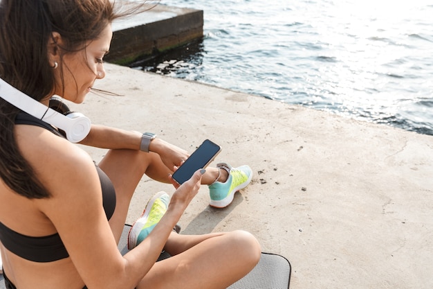 Vista dall'alto di una giovane donna attraente fitness utilizzando il telefono cellulare mentre si riposa in spiaggia dopo il jogging