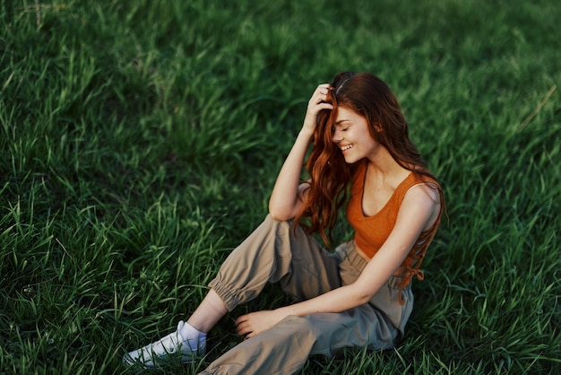 Vista dall'alto di una giovane donna dai capelli rossi seduta nella natura sull'erba verde e sorridente e coprendosi il viso con i capelli per imbarazzo