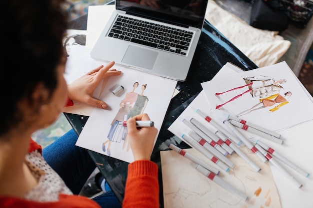 Vista dall'alto di una giovane donna seduta al tavolo con un laptop che disegna sognante illustrazioni di moda mentre trascorre del tempo in un'officina moderna