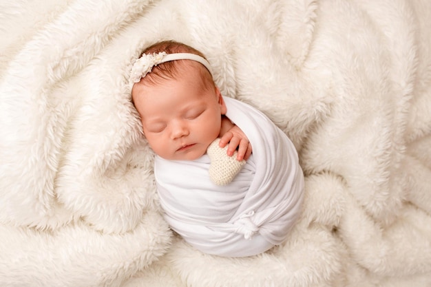 Vista dall'alto di una neonata che dorme in un bozzolo bianco con una benda bianca e un fiore in testa su uno sfondo bianco Bel ritratto di una bambina 7 giorni una settimana Macrofotografia