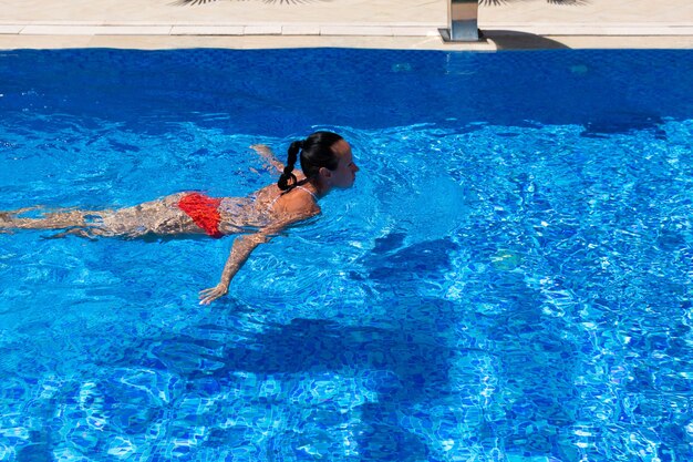 Vista dall'alto di una ragazza abbronzata, femmina, modella in costume da bagno rosso, che nuota nell'acqua blu della piscina.