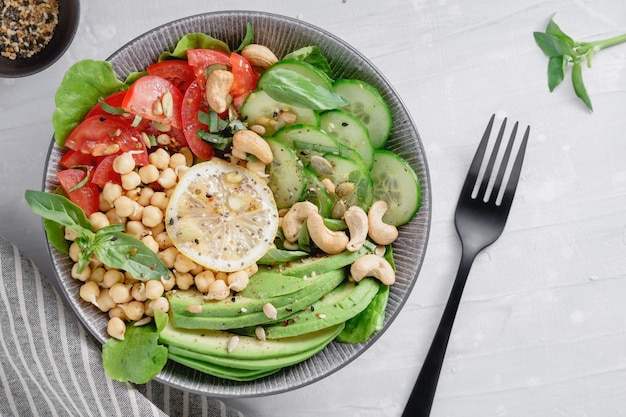 Vista dall'alto di una sana ciotola vegana con avocado crudo ceci pomodori anacardi cetriolo verdi e semi su un tavolo da cucina bianco